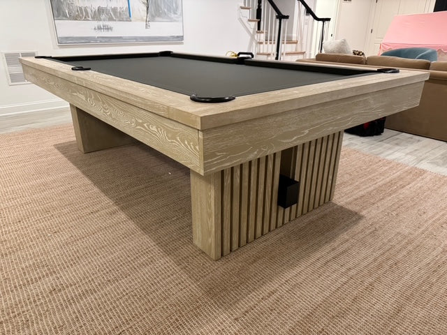 Wooden pool table with a black felt surface in a room with a staircase and furniture.